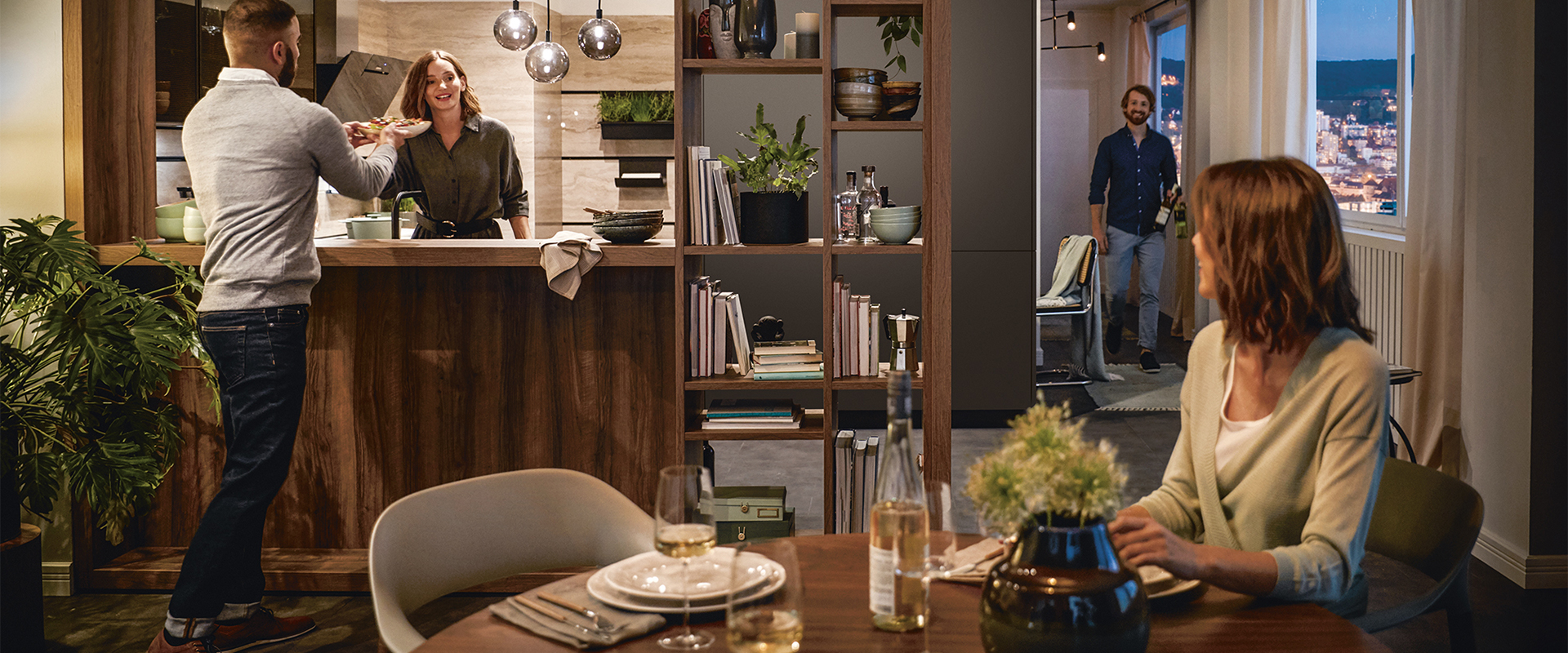 Gesellige&#x20;Szene&#x20;in&#x20;einer&#x20;modernen&#x20;K&#xFC;che&#x3A;&#x20;Ein&#x20;Mann&#x20;reicht&#x20;einer&#x20;Frau&#x20;ein&#x20;Gericht&#x20;&#xFC;ber&#x20;eine&#x20;K&#xFC;cheninsel,&#x20;w&#xE4;hrend&#x20;ein&#x20;weiterer&#x20;Gast&#x20;mit&#x20;Weinflaschen&#x20;hereinkommt.&#x20;Eine&#x20;Frau&#x20;sitzt&#x20;am&#x20;gedeckten&#x20;Esstisch.