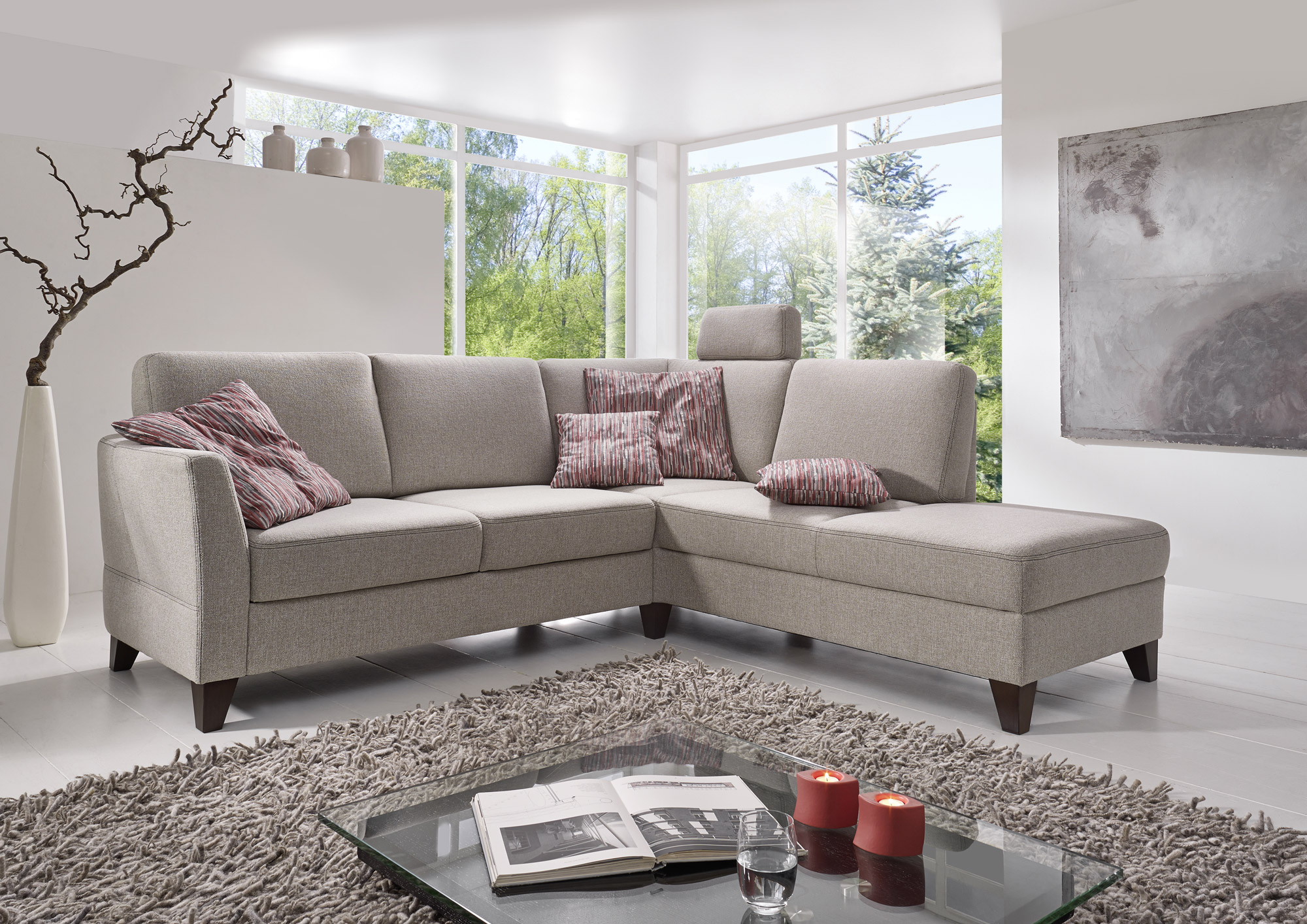 Beiges&#x20;Ecksofa&#x20;mit&#x20;roten&#x20;Zierkissen&#x20;vor&#x20;gro&#xDF;en&#x20;Fenstern,&#x20;daneben&#x20;ein&#x20;Teppich&#x20;und&#x20;eine&#x20;moderne&#x20;Vase&#x20;mit&#x20;Zweig.