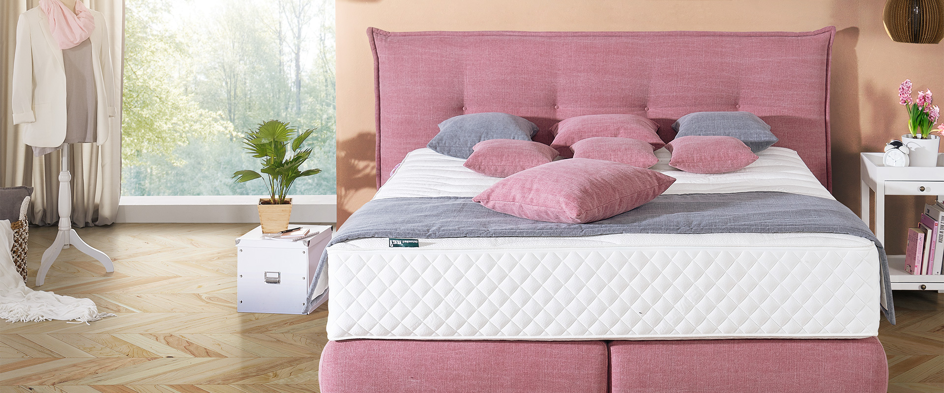 Schlafzimmer&#x20;mit&#x20;rosafarbenem&#x20;Polsterbett,&#x20;grauer&#x20;Bettdecke&#x20;und&#x20;mehreren&#x20;rosa&#x20;und&#x20;grauen&#x20;Kissen,&#x20;daneben&#x20;ein&#x20;Nachttisch&#x20;mit&#x20;Pflanze&#x20;und&#x20;Wecker.