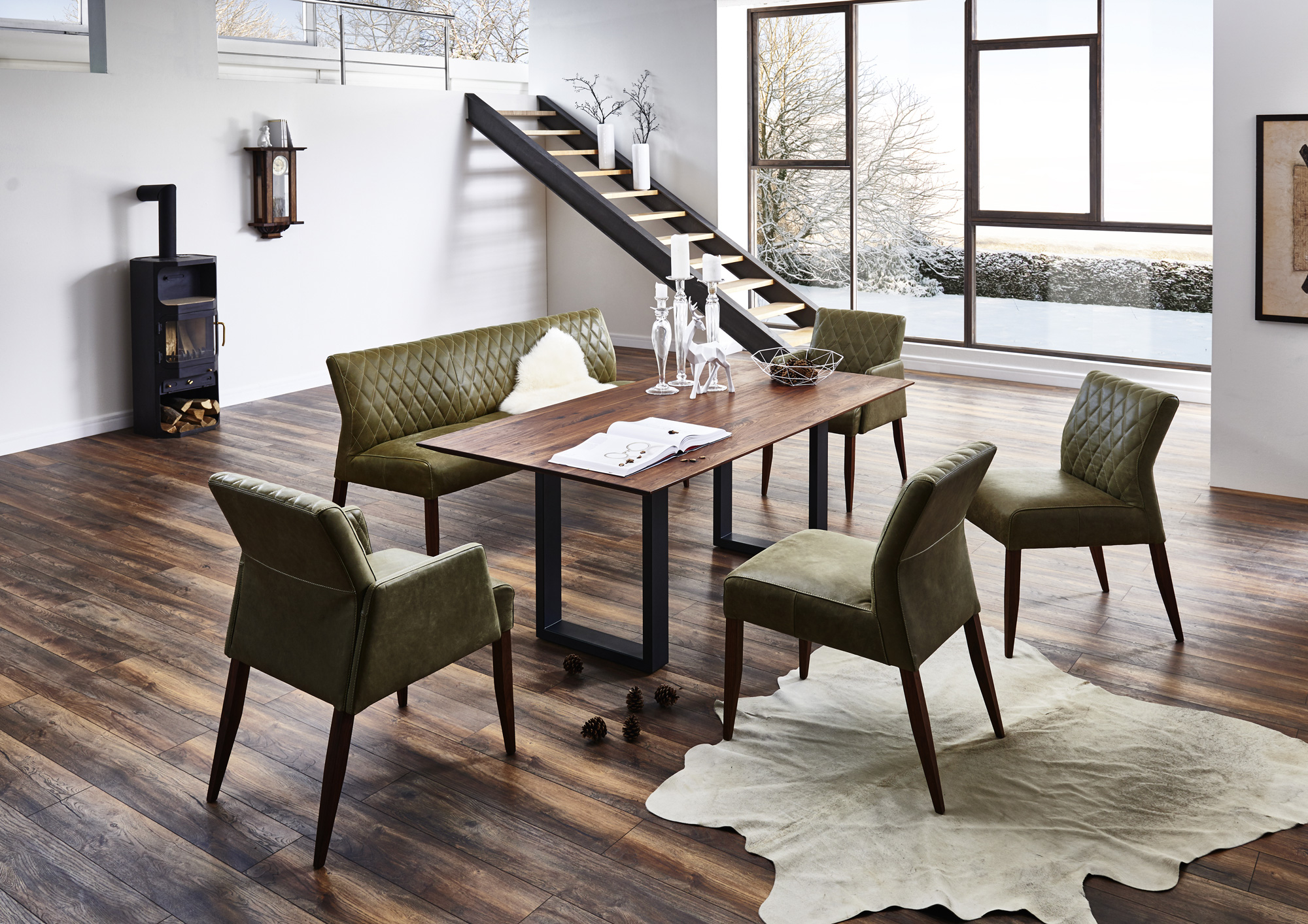 Ein&#x20;Esszimmer&#x20;mit&#x20;modernem&#x20;Design&#x3A;&#x20;Holztisch,&#x20;gr&#xFC;ne&#x20;Polsterst&#xFC;hle&#x20;und&#x20;-bank&#x20;auf&#x20;dunklem&#x20;Holzboden,&#x20;helles&#x20;Tageslicht&#x20;durch&#x20;gro&#xDF;e&#x20;Fenster,&#x20;Holzofen&#x20;in&#x20;der&#x20;Ecke.