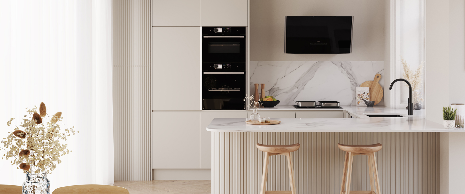 Moderne&#x20;K&#xFC;che&#x20;in&#x20;Cremefarben&#x20;mit&#x20;Marmorarbeitsplatte,&#x20;integriertem&#x20;Backofen&#x20;und&#x20;schwarzer&#x20;Dunstabzugshaube.&#x20;Zwei&#x20;Holzbarhocker&#x20;stehen&#x20;an&#x20;der&#x20;K&#xFC;cheninsel,&#x20;dekoriert&#x20;mit&#x20;Trockenblumen&#x20;und&#x20;Schalen.