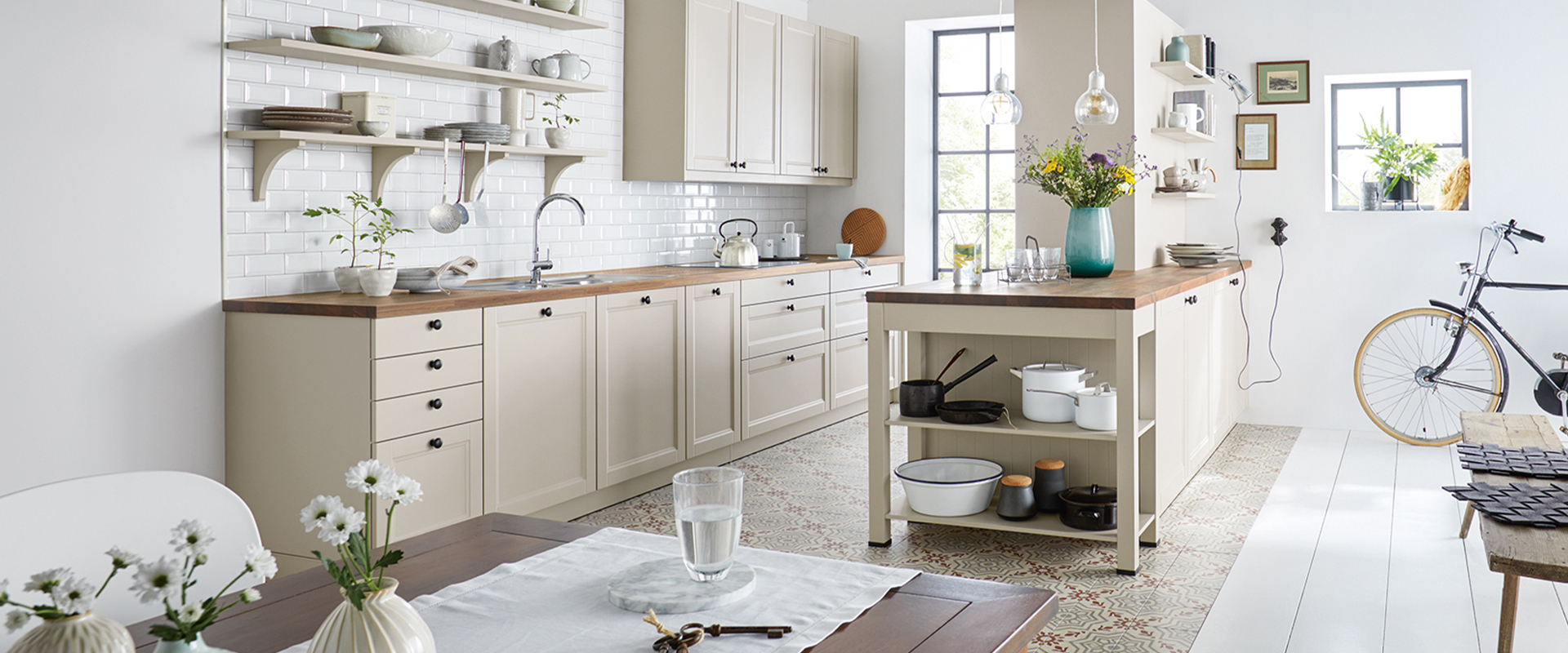 Helle&#x20;K&#xFC;che&#x20;im&#x20;Landhausstil&#x20;mit&#x20;cremefarbenen&#x20;Schr&#xE4;nken,&#x20;Holztresen&#x20;und&#x20;offener&#x20;Regaleinrichtung.&#x20;Im&#x20;Hintergrund&#x20;ist&#x20;ein&#x20;Fahrrad&#x20;und&#x20;ein&#x20;kleines&#x20;Fenster&#x20;mit&#x20;Pflanzen&#x20;zu&#x20;sehen.