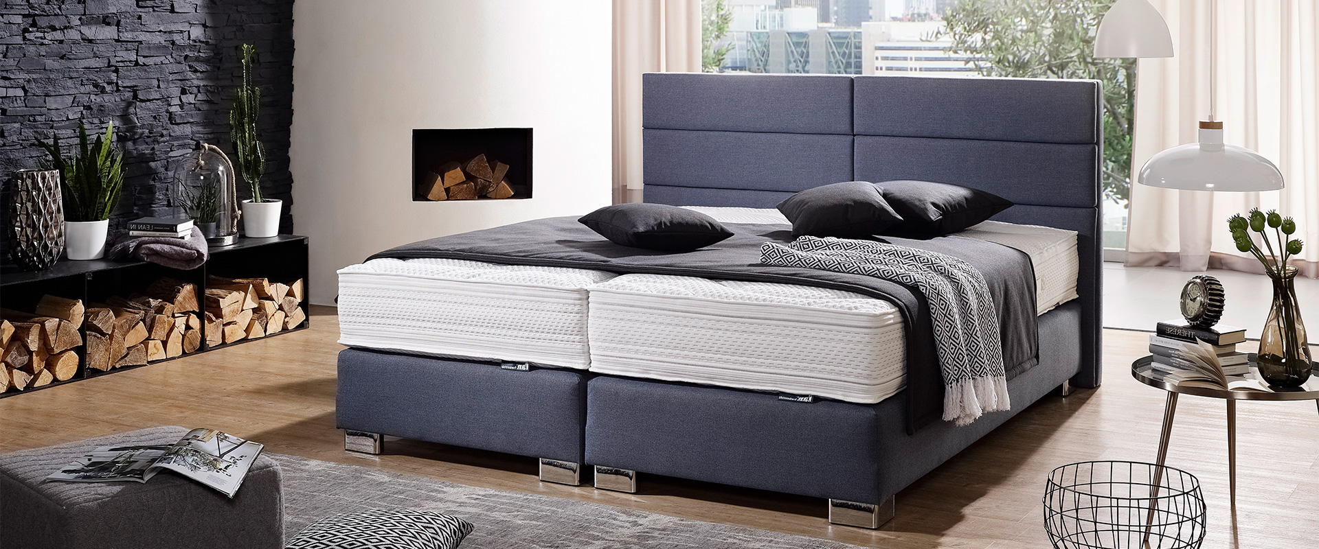 Modernes&#x20;Schlafzimmer&#x20;mit&#x20;dunkelblauem&#x20;Boxspringbett,&#x20;schwarzen&#x20;Kissen&#x20;und&#x20;gemusterter&#x20;Decke.&#x20;Eine&#x20;Steinwand&#x20;mit&#x20;Regalen,&#x20;Holzlager&#x20;und&#x20;Pflanzen&#x20;sorgt&#x20;f&#xFC;r&#x20;einen&#x20;nat&#xFC;rlichen&#x20;Akzent.