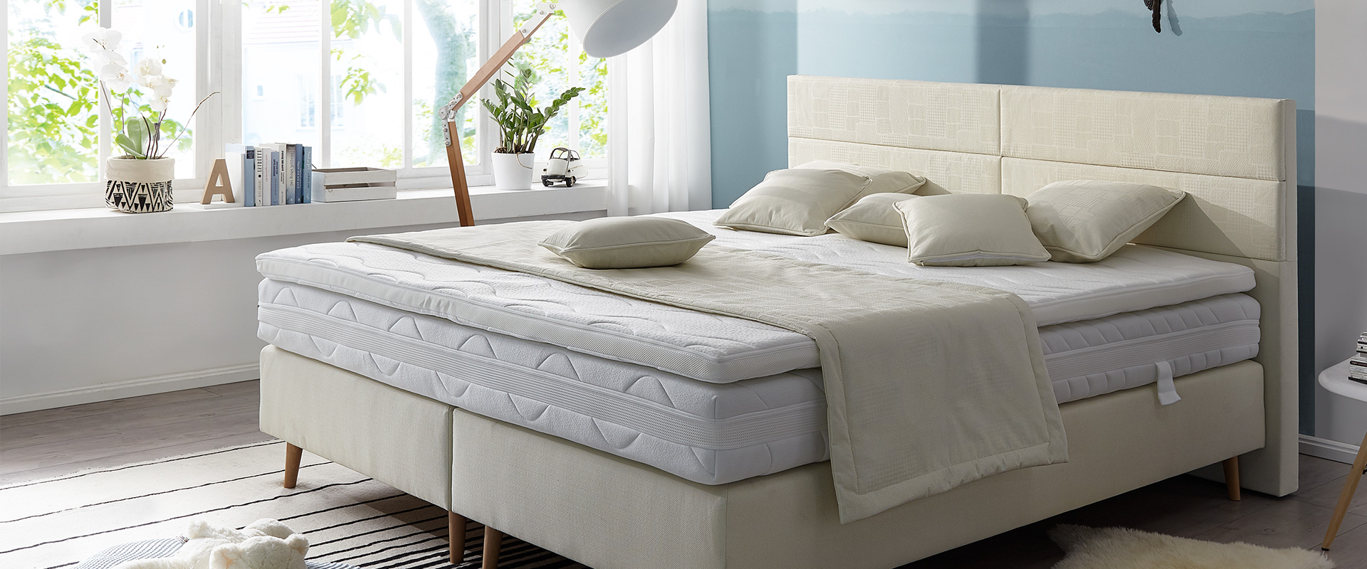 Helles&#x20;Schlafzimmer&#x20;mit&#x20;gepolstertem&#x20;Bett,&#x20;mehreren&#x20;Kissen&#x20;und&#x20;einer&#x20;gro&#xDF;en&#x20;Stehlampe.&#x20;Auf&#x20;der&#x20;Fensterbank&#x20;stehen&#x20;B&#xFC;cher&#x20;und&#x20;Pflanzen,&#x20;der&#x20;Raum&#x20;ist&#x20;in&#x20;sanften&#x20;T&#xF6;nen&#x20;gehalten.