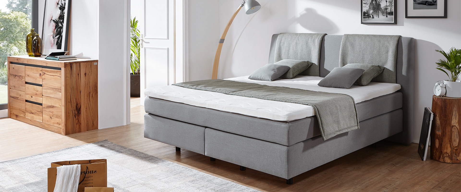 Helles&#x20;Schlafzimmer&#x20;mit&#x20;grauem&#x20;Boxspringbett,&#x20;Holzkommode&#x20;und&#x20;Stehlampe.&#x20;Dekoriert&#x20;mit&#x20;Kissen,&#x20;Pflanzen&#x20;und&#x20;Wandbildern&#x20;f&#xFC;r&#x20;eine&#x20;gem&#xFC;tliche&#x20;Atmosph&#xE4;re.