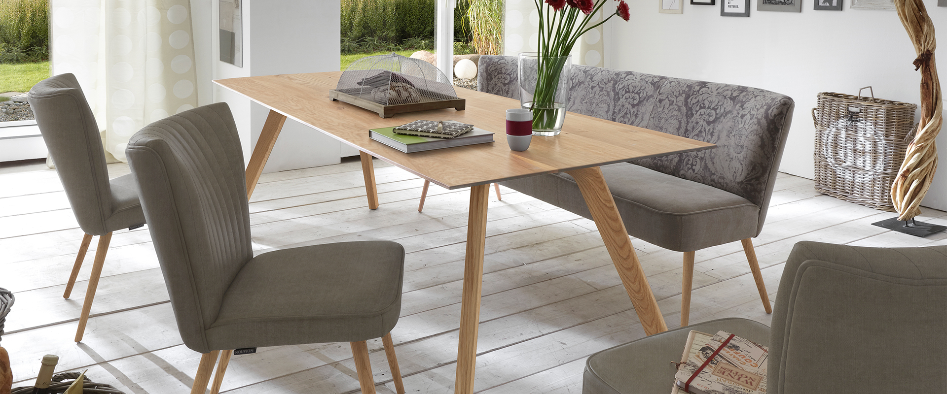 Esszimmer&#x20;mit&#x20;hellem&#x20;Holztisch,&#x20;grauen&#x20;Polsterst&#xFC;hlen&#x20;und&#x20;-bank,&#x20;dekoriert&#x20;mit&#x20;Blumen&#x20;und&#x20;B&#xFC;chern.&#x20;Gro&#xDF;e&#x20;Fenster&#x20;bieten&#x20;Blick&#x20;ins&#x20;Gr&#xFC;ne.