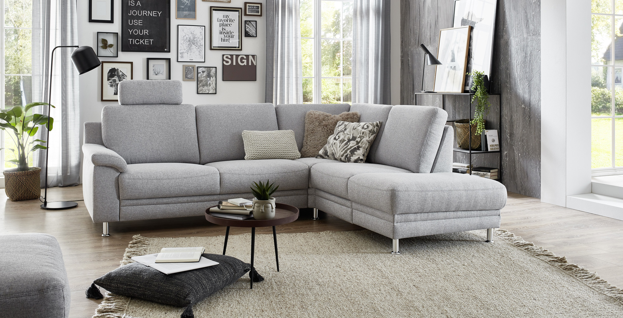 Heller&#x20;Wohnraum&#x20;mit&#x20;grauem&#x20;Ecksofa,&#x20;dekorativen&#x20;Kissen,&#x20;kleiner&#x20;Holztisch&#x20;auf&#x20;Teppich,&#x20;Bilderwand&#x20;und&#x20;gro&#xDF;en&#x20;Fenstern&#x20;mit&#x20;Vorh&#xE4;ngen