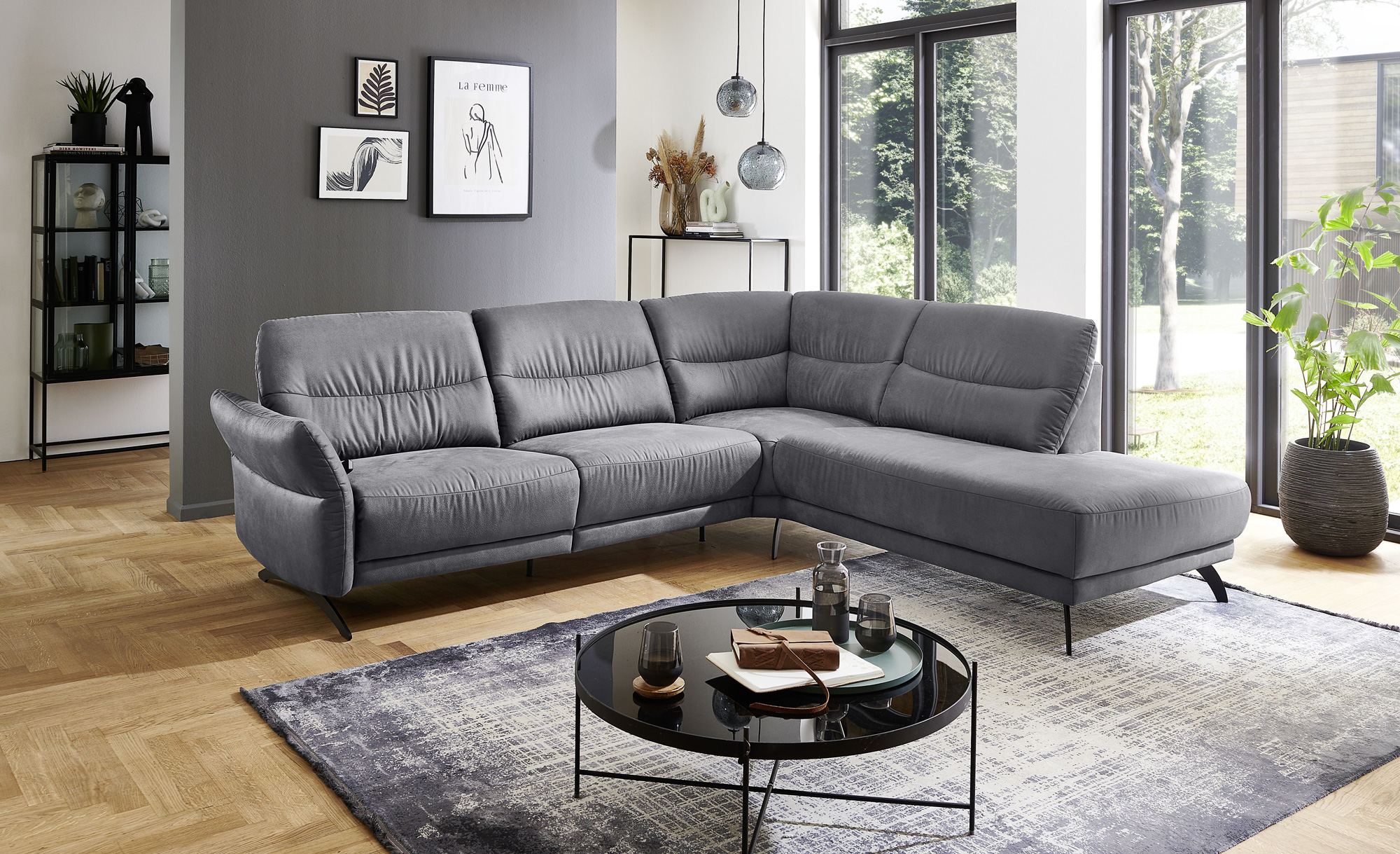 Wohnzimmer-Set&#x20;aus&#xA0;&#x20;grauem&#x20;Ecksofa,&#x20;rundem&#x20;Couchtisch,&#x20;Teppich,&#x20;gro&#xDF;en&#x20;Fenstern&#x20;und&#x20;dekorativem&#x20;Regal&#x20;vor&#x20;einer&#x20;grauen&#x20;Wand